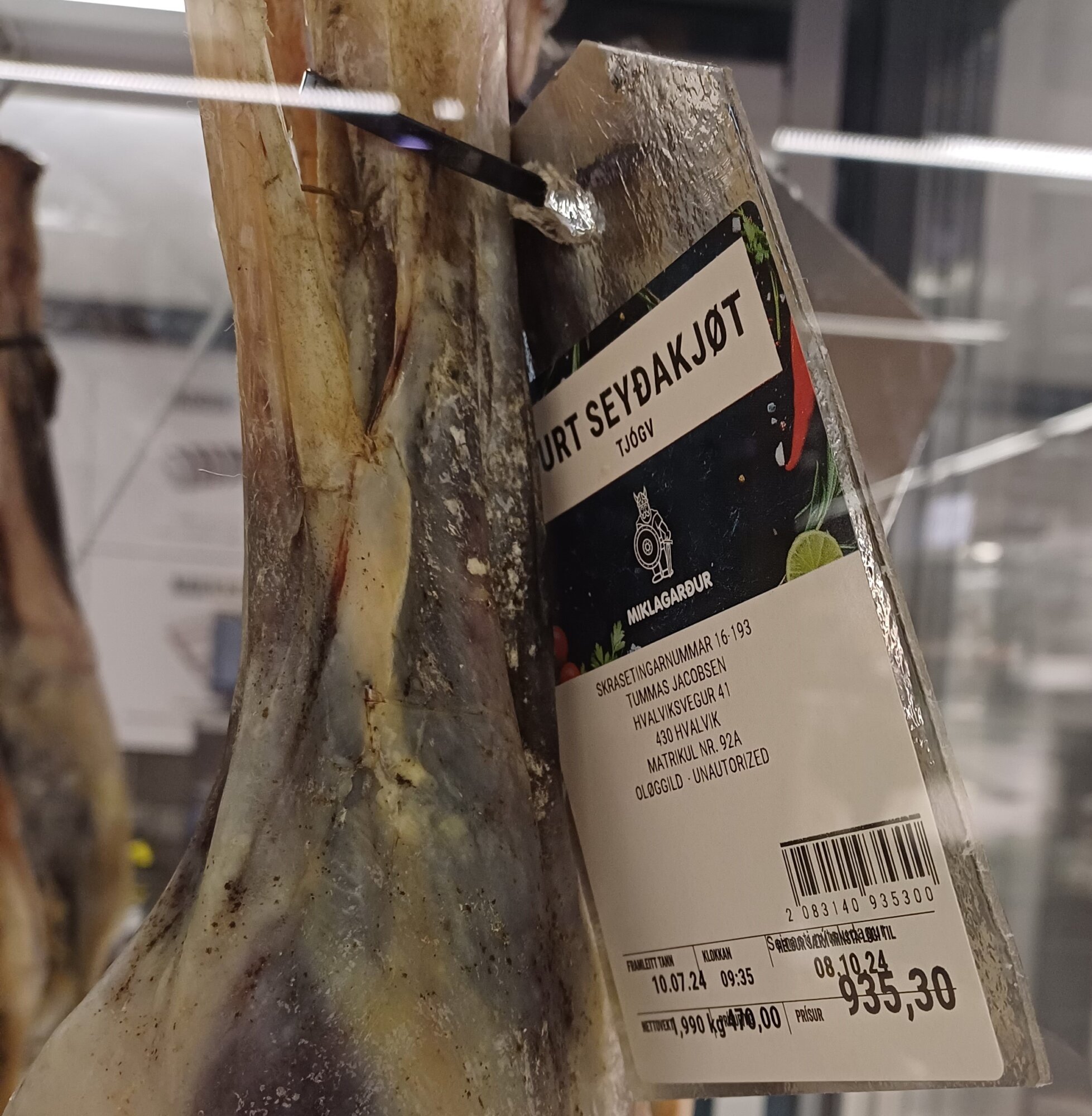 Wisząca w chłodni suszona tradycyjna udka jagnięca (Skerpikjøt / Seyðakjøt) z przypiętą etykietą sklepu Miklagarður, wagą 1,990 kg i ceną 935,30 DKK