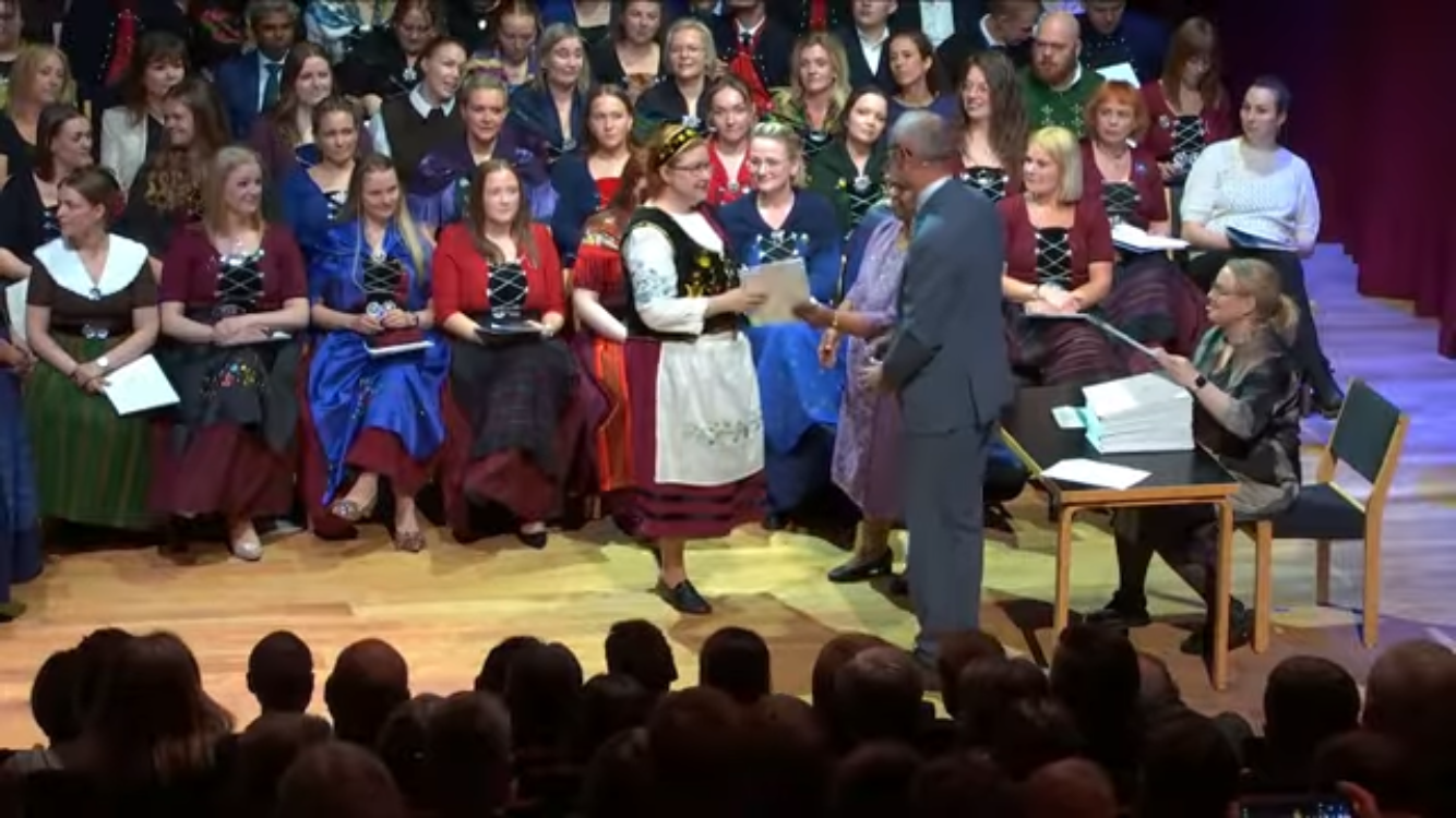 Oficjalna ceremonia wręczenia dyplomów ukończenia studiów na Wyspach Owczych. Absolwenci w tradycyjnych strojach odbierają dokumenty na scenie.